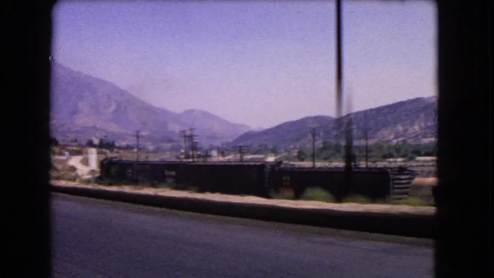 1966 California roadside view from a moving car, showing dark freight rail cars, utility poles, dry hills, and mountains.