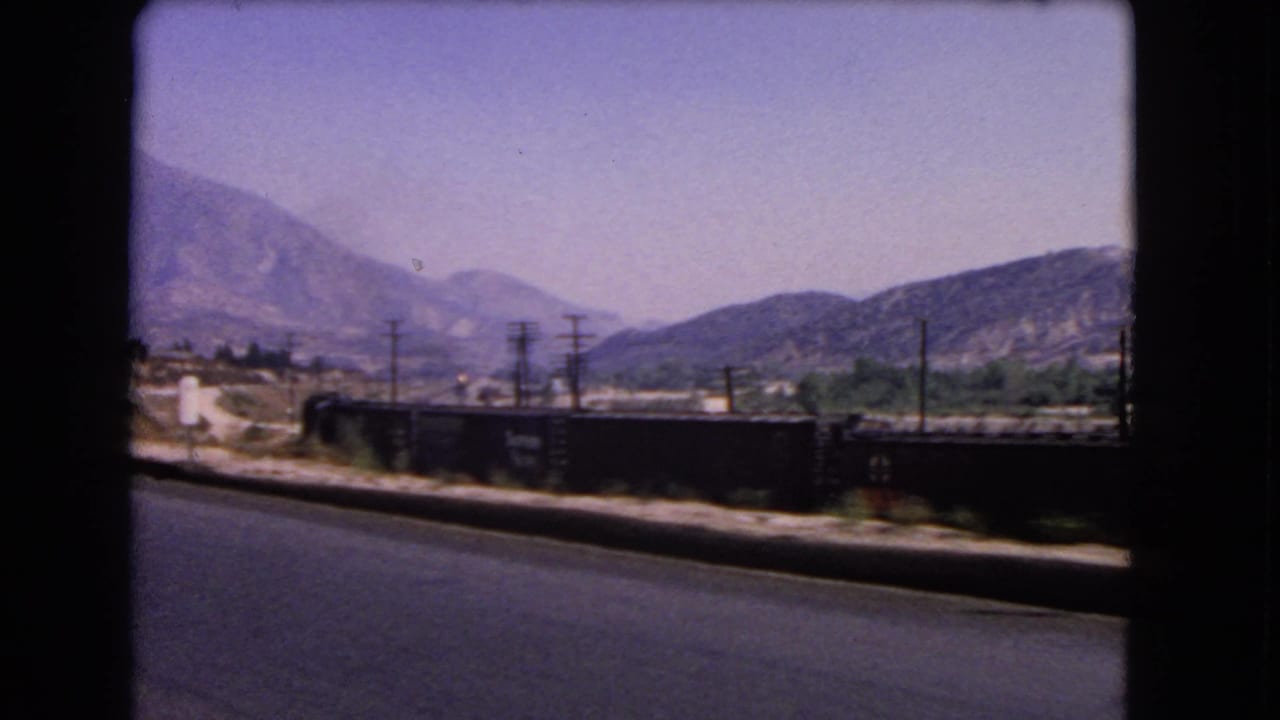 1966 California 8mm frame with a moving roadside view of rail cars, utility poles, dry terrain, and distant mountains.
