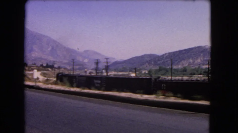 1966 California 8mm frame with a moving roadside view of rail cars, utility poles, dry terrain, and distant mountains.