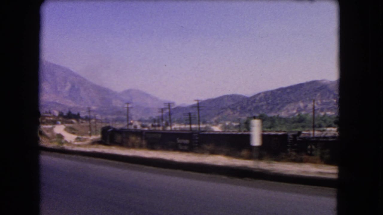1966 California roadside frame showing rail cars, utility poles, a signpost, dry hills, and layered mountain ridges.