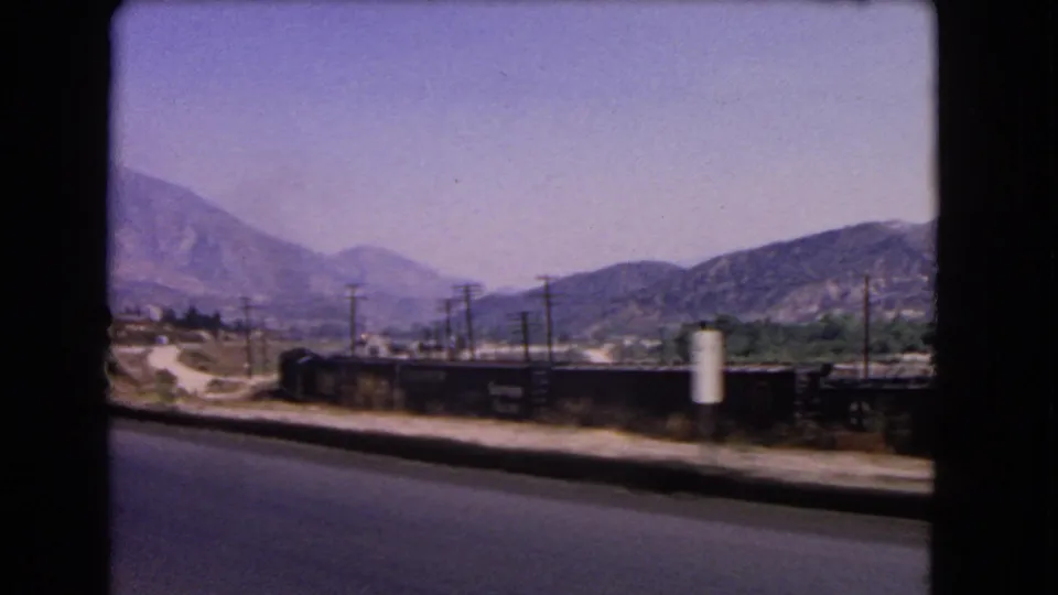 1966 California roadside frame showing rail cars, utility poles, a signpost, dry hills, and layered mountain ridges.