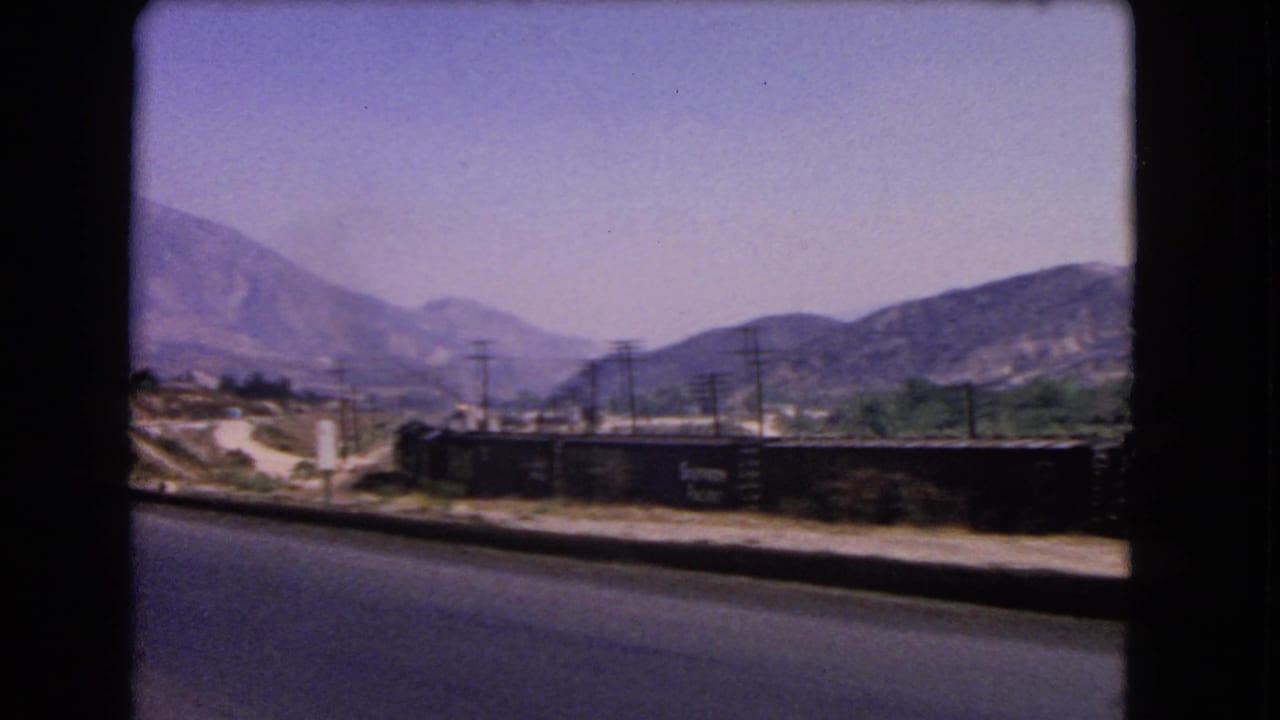 1966 California 8mm frame showing a paved road beside a freight train, utility poles, dry ground, and mountains in haze.