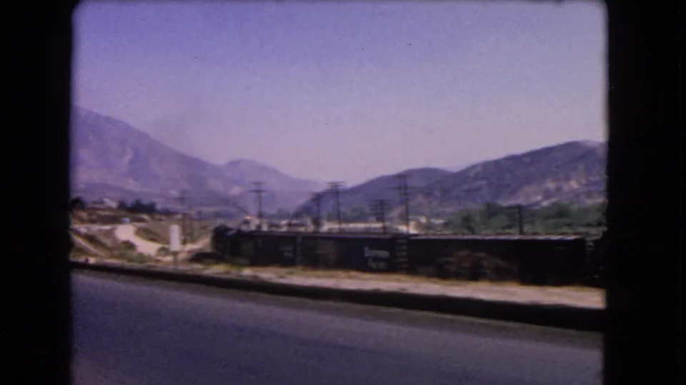 1966 California 8mm frame showing a paved road beside a freight train, utility poles, dry ground, and mountains in haze.