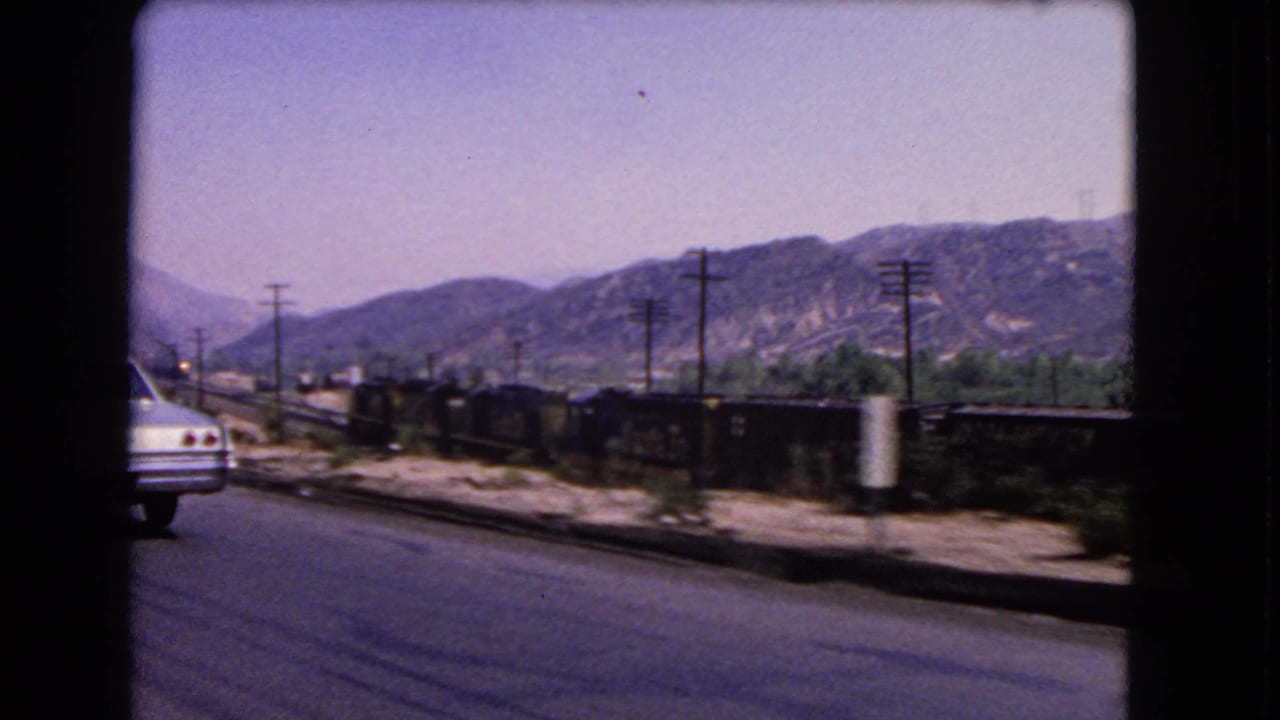Moving-car view in California, 1966, with a white car ahead, parallel freight train, utility poles, and mountains beyond.