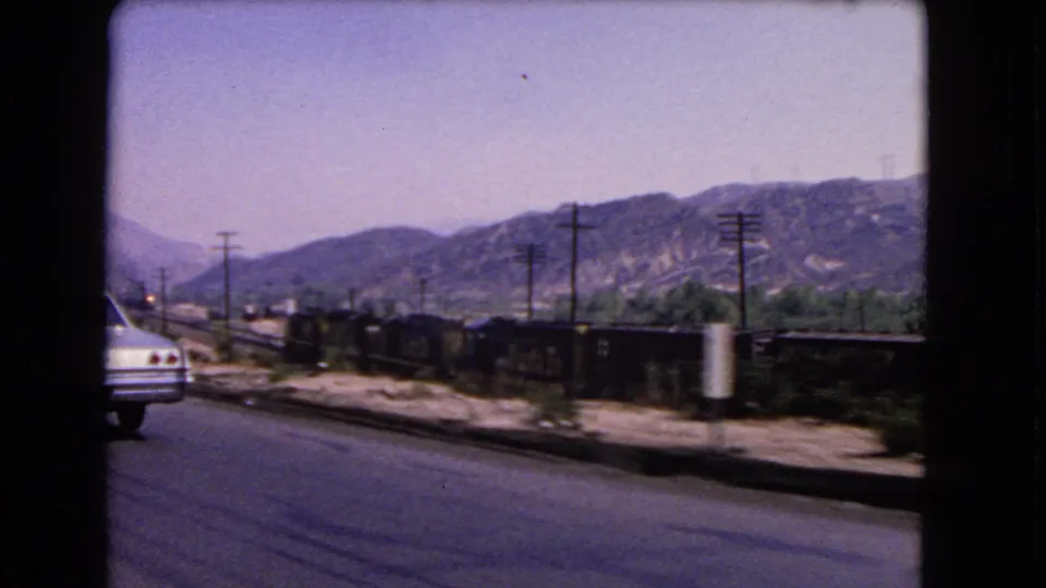 Moving-car view in California, 1966, with a white car ahead, parallel freight train, utility poles, and mountains beyond.