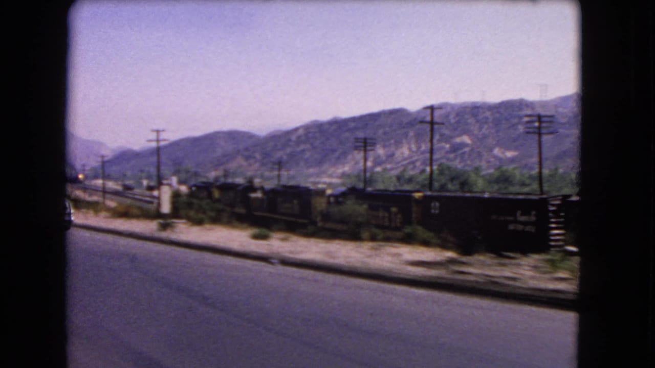 1966 California road footage showing dark freight cars, roadside poles, dry shoulder, and a mountain range under pale sky.