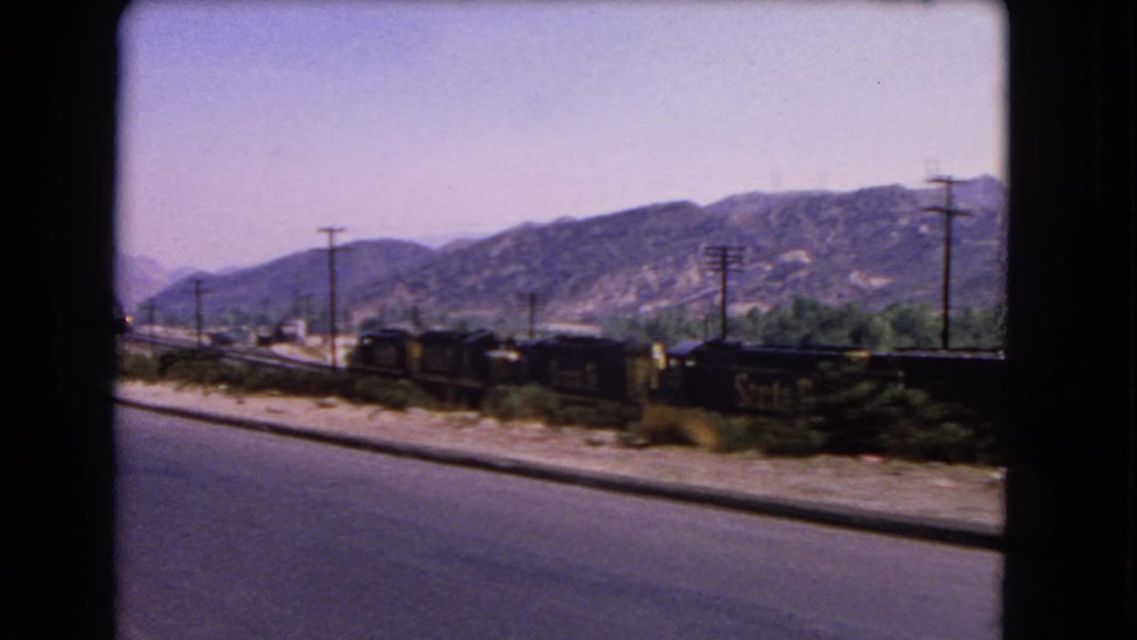 Passenger-view 8mm frame from 1966 California, showing road pavement, rail cars, utility poles, shrubs, and distant mountains.