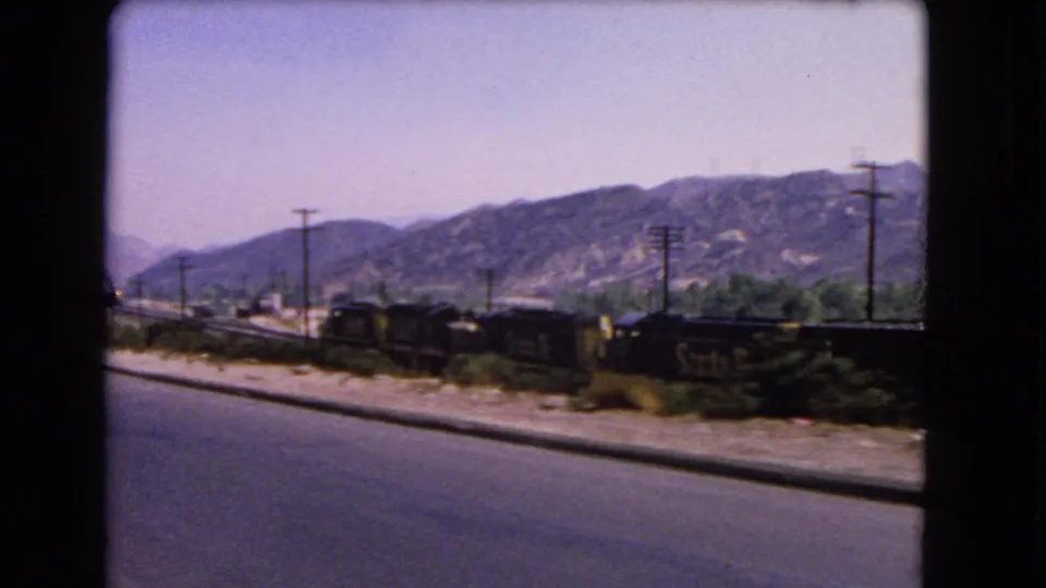 Passenger-view 8mm frame from 1966 California, showing road pavement, rail cars, utility poles, shrubs, and distant mountains.