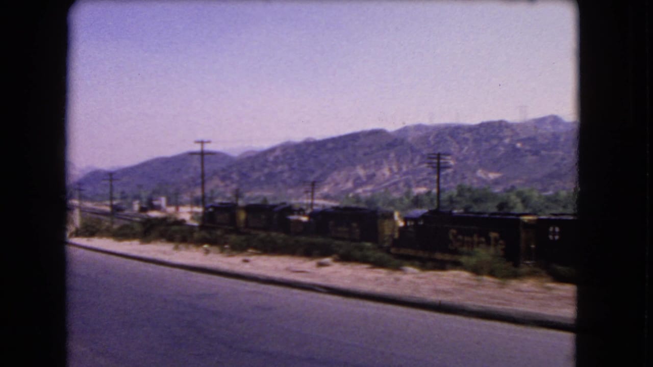 1966 California home-movie view of a Santa Fe freight train passing beside a road, with dry hills and utility poles beyond.