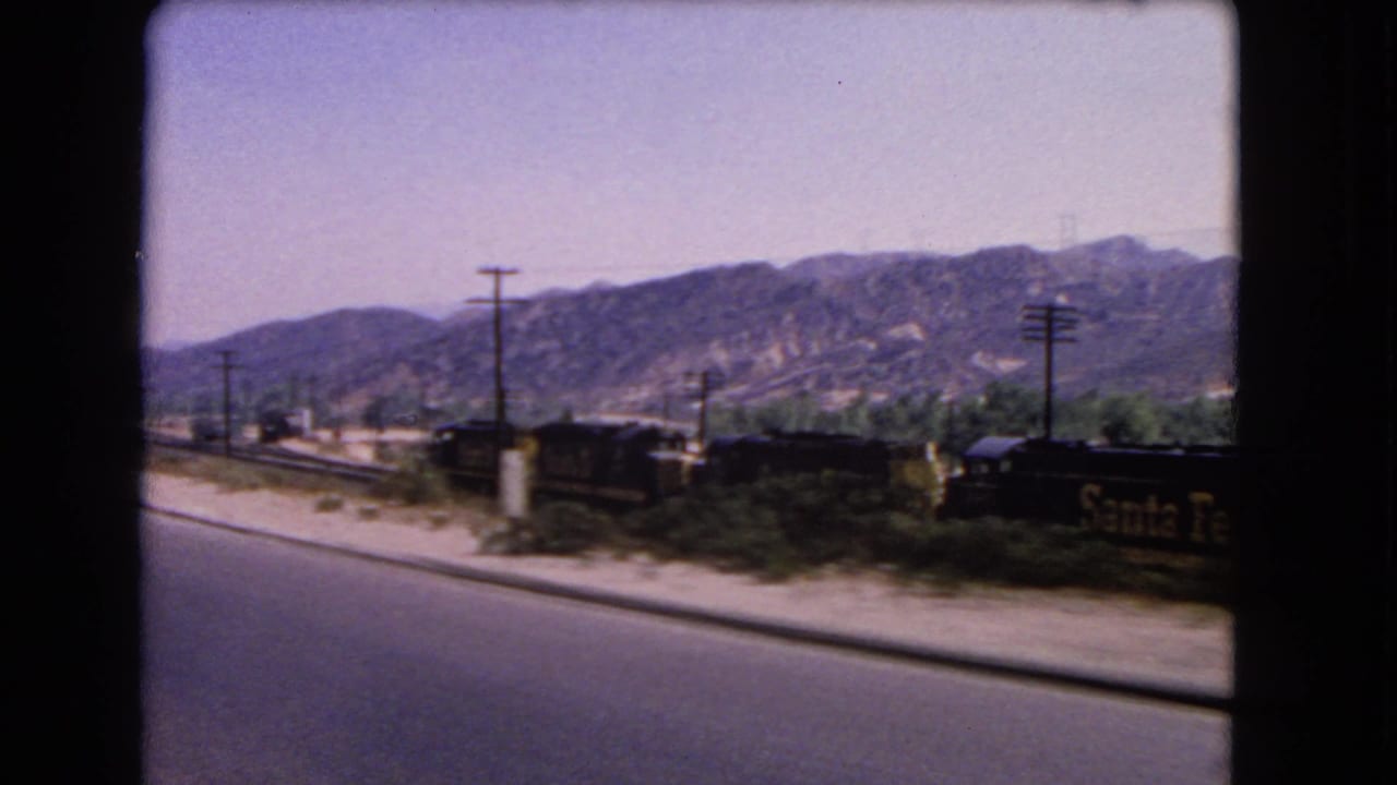 Freight cars pass along tracks near a road, with Santa Fe lettering and hills behind.