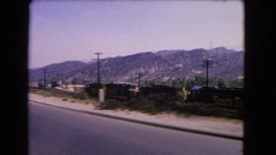 Santa Fe freight cars pass through a sunlit California roadside scene, framed by a vehicle or train window.