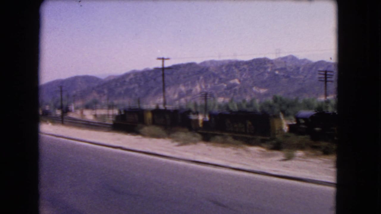 Motion-blurred 1966 California view of freight cars beside a road, with utility poles and dry hills in the distance.