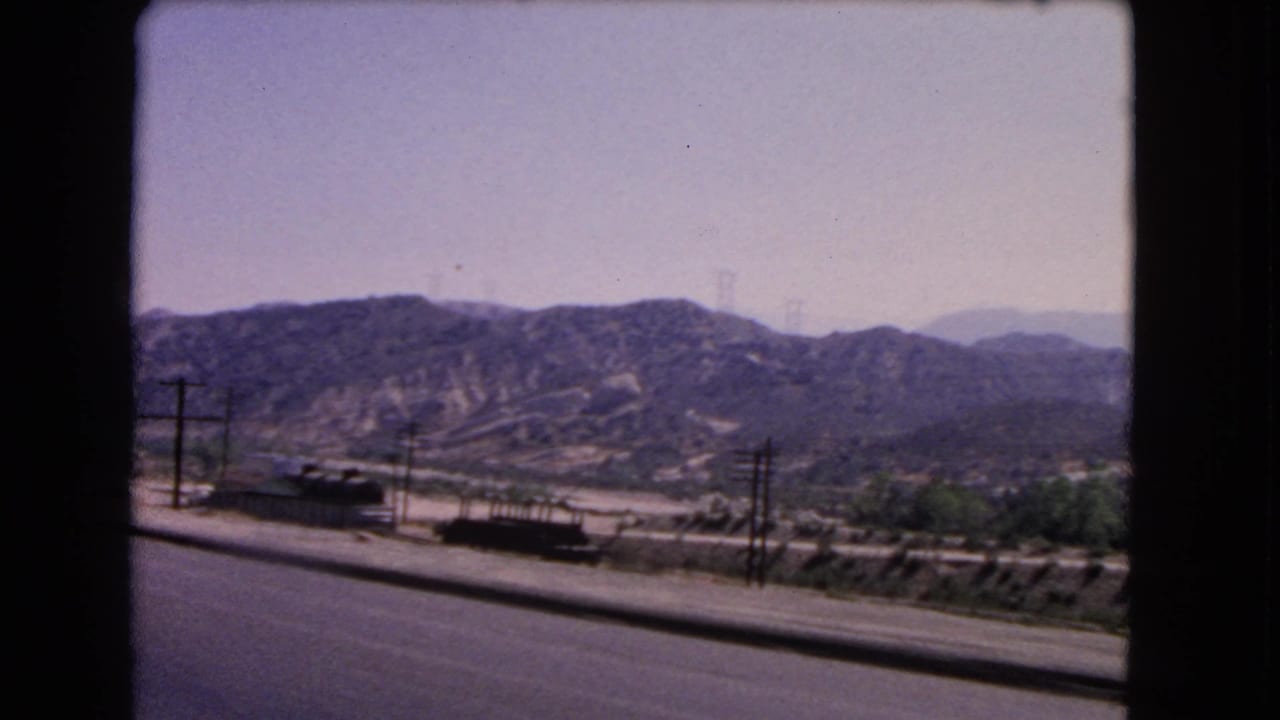Wider California home-movie view of freight rail activity near a road, with low hills and utility poles across the landscape.