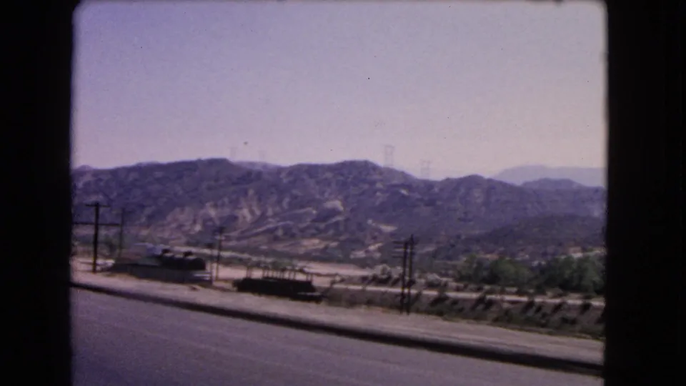 Wider California home-movie view of freight rail activity near a road, with low hills and utility poles across the landscape.