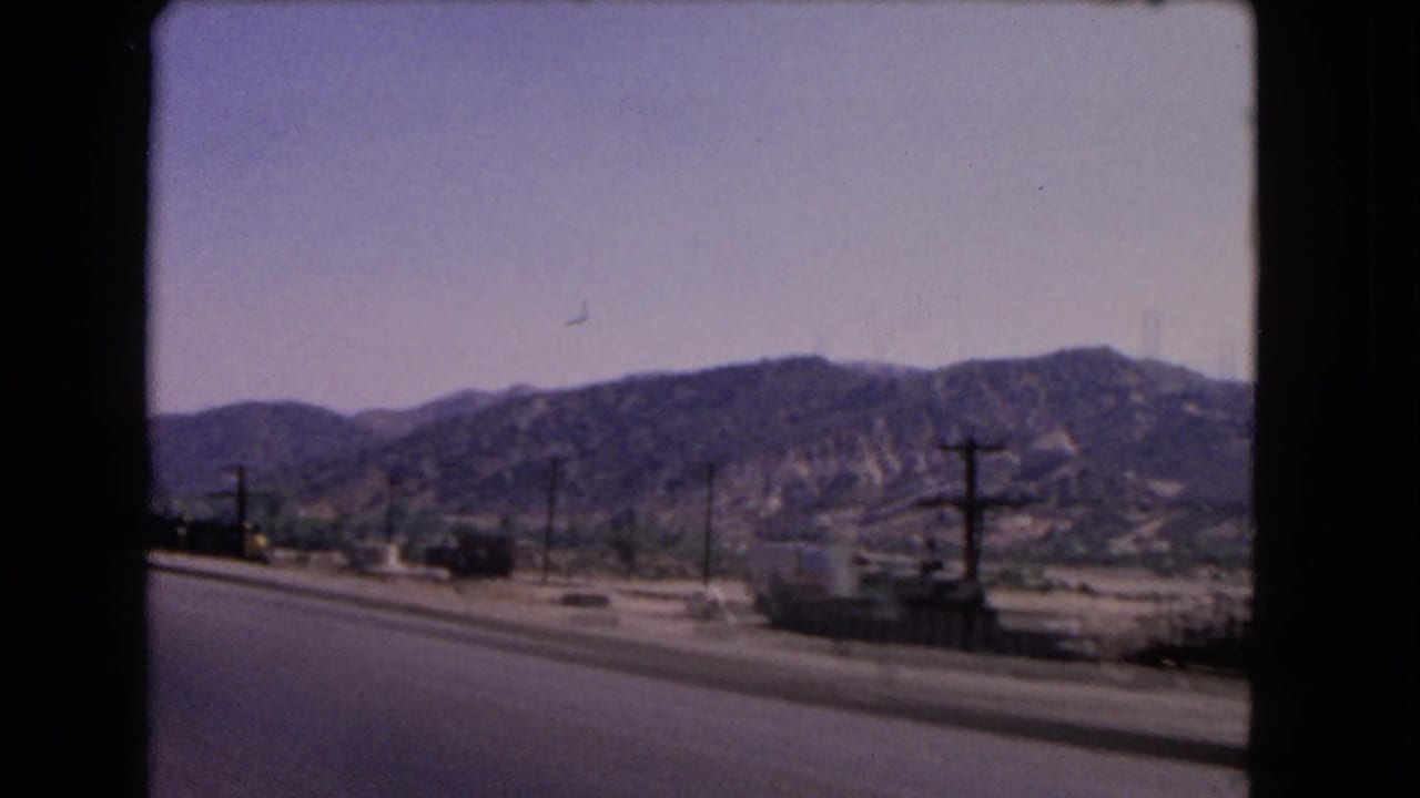 1966 California 8mm roadside view with dry hills, utility poles, low buildings, and a paved road in the foreground.