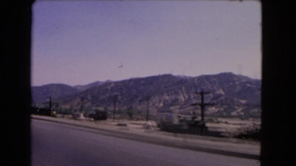 1966 California 8mm roadside view with dry hills, utility poles, low buildings, and a paved road in the foreground.