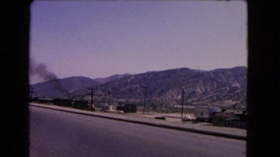 1966 California roadside scene with a smoke-producing train, utility poles, cars, and dry hills beyond the road.
