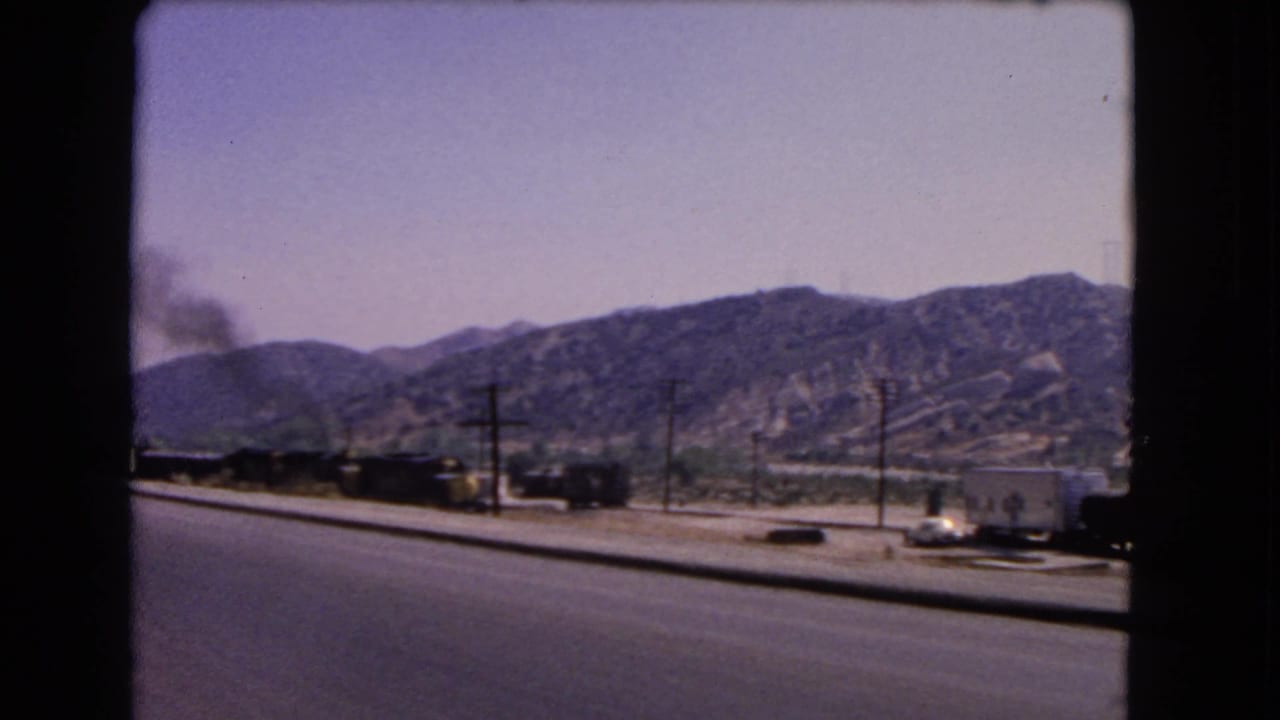 1966 California 8mm frame showing train cars with smoke, a roadside foreground, utility poles, and dry hillside terrain.