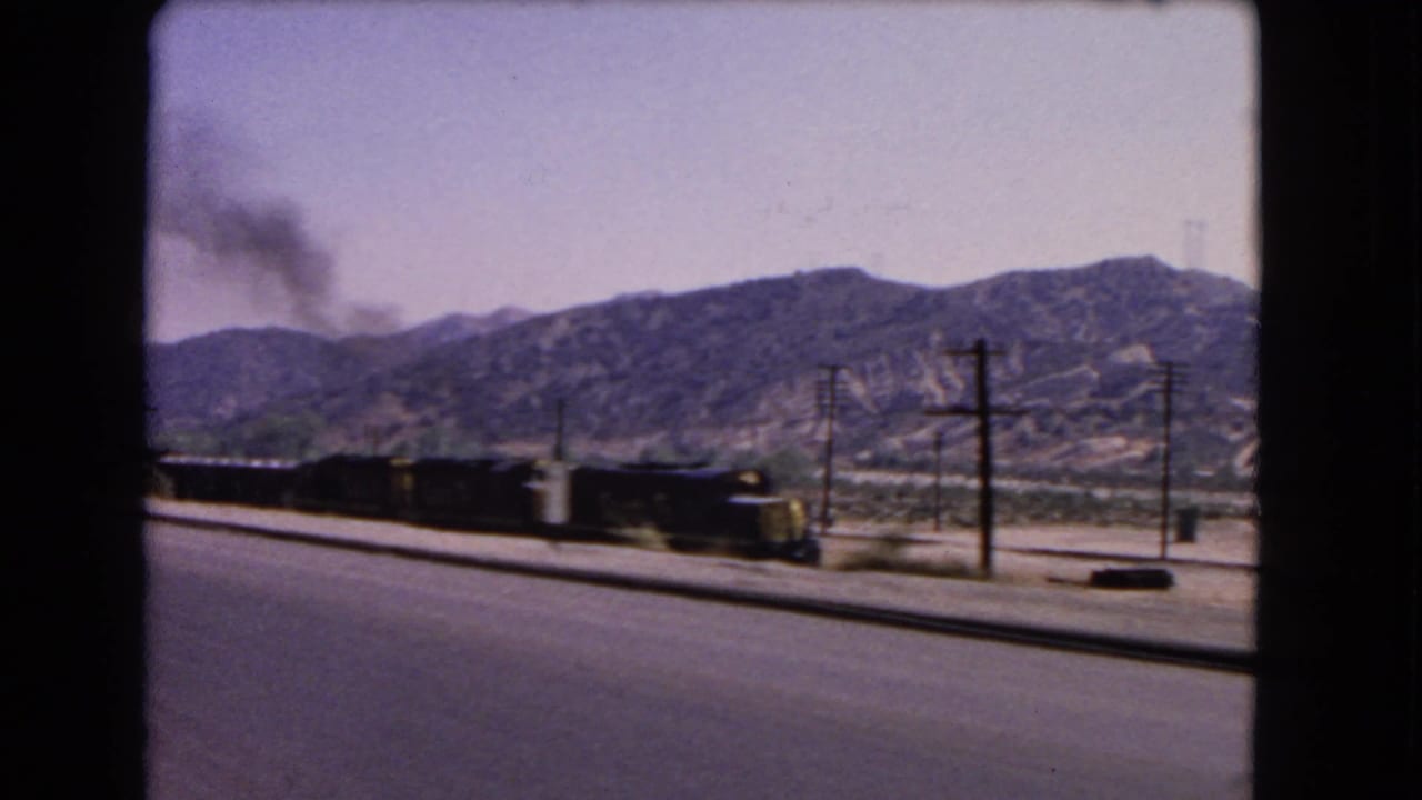 1966 California home-movie frame showing a freight train moving beside a paved road below dry mountain slopes.