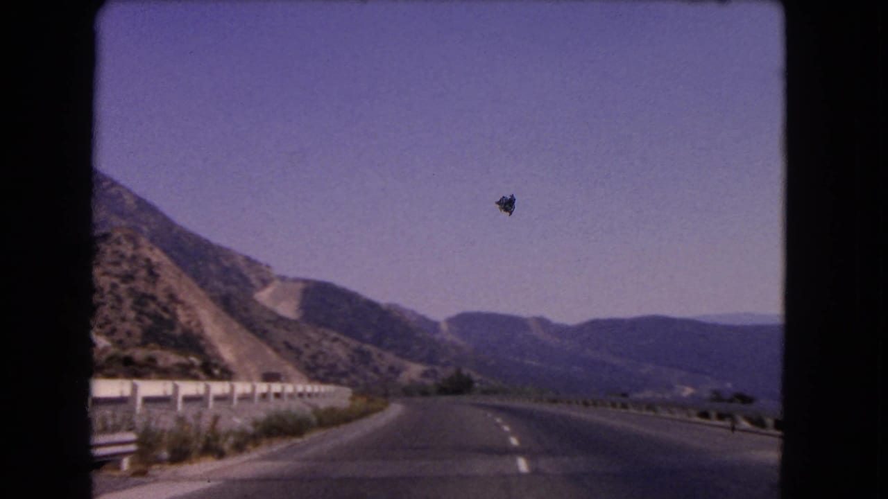 View from a moving vehicle on a California mountain highway, with guardrail, open road, and dry hills under clear sky.