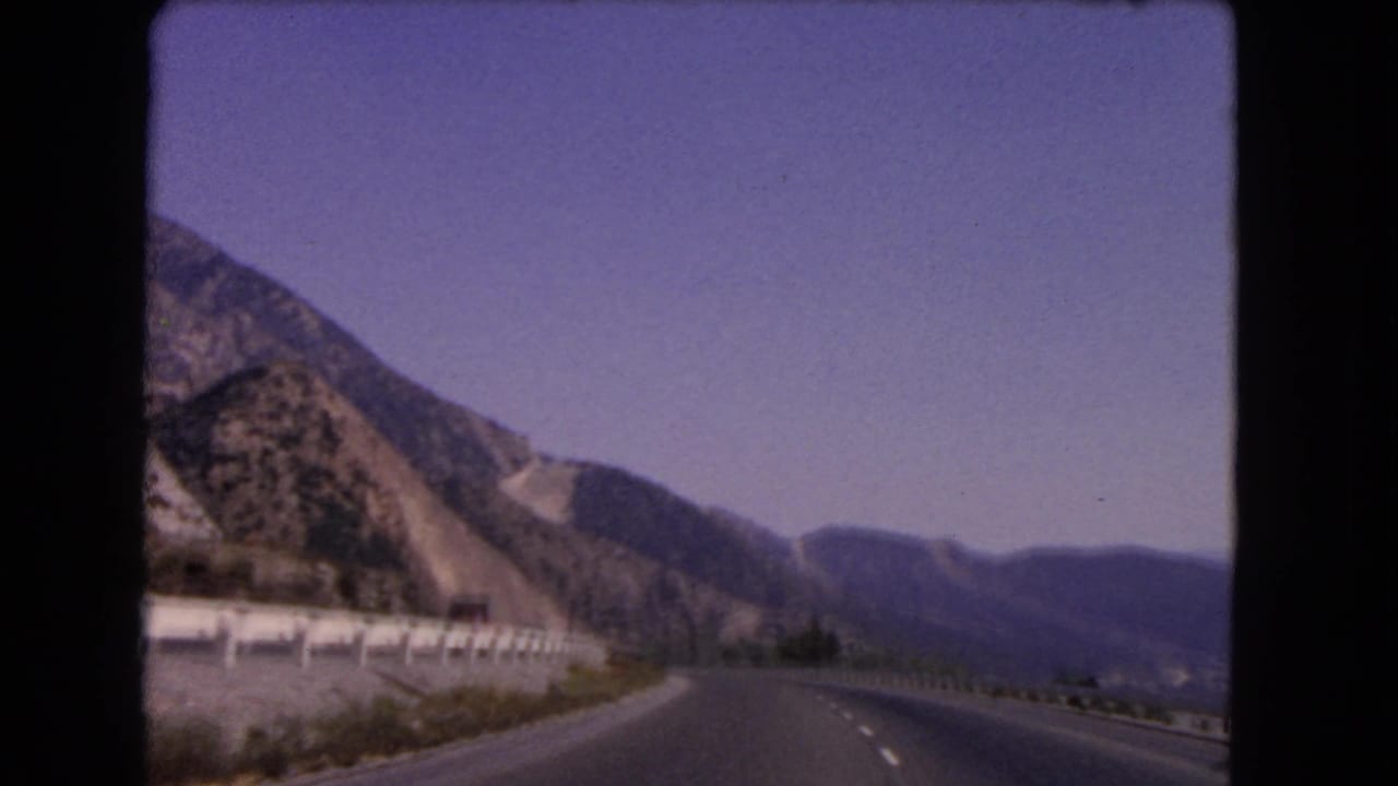 A 1966 California road-trip frame shows a curving highway, roadside guardrail, and rugged mountain slopes from a moving car.