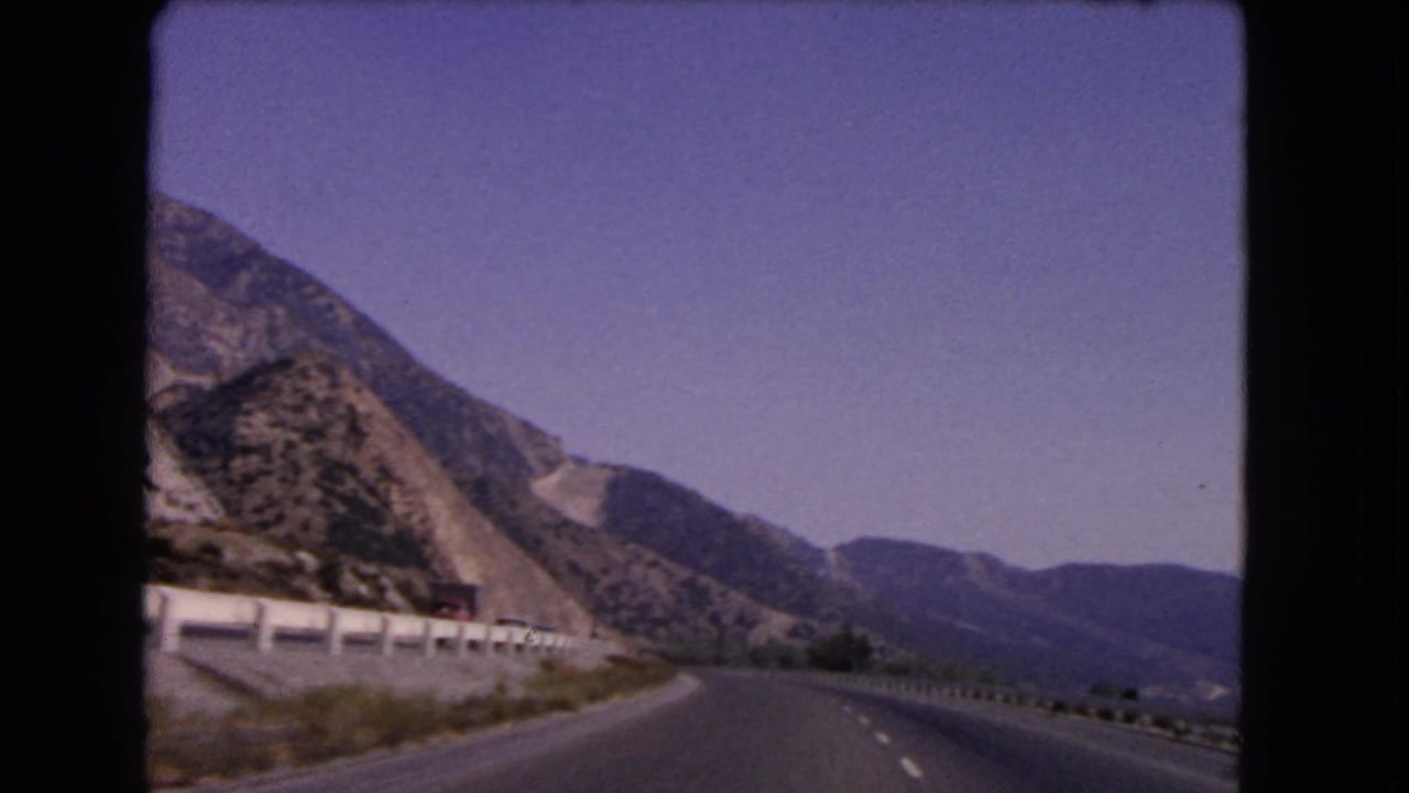1966 California 8mm view from a moving car on a curving mountain highway bordered by a white guardrail and rocky slopes.