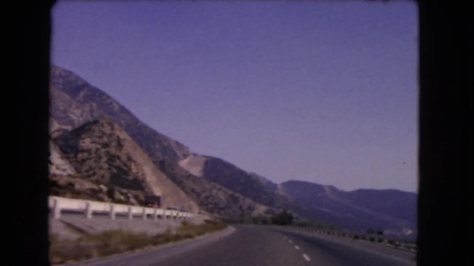 1966 California 8mm view from a moving car on a curving mountain highway bordered by a white guardrail and rocky slopes.