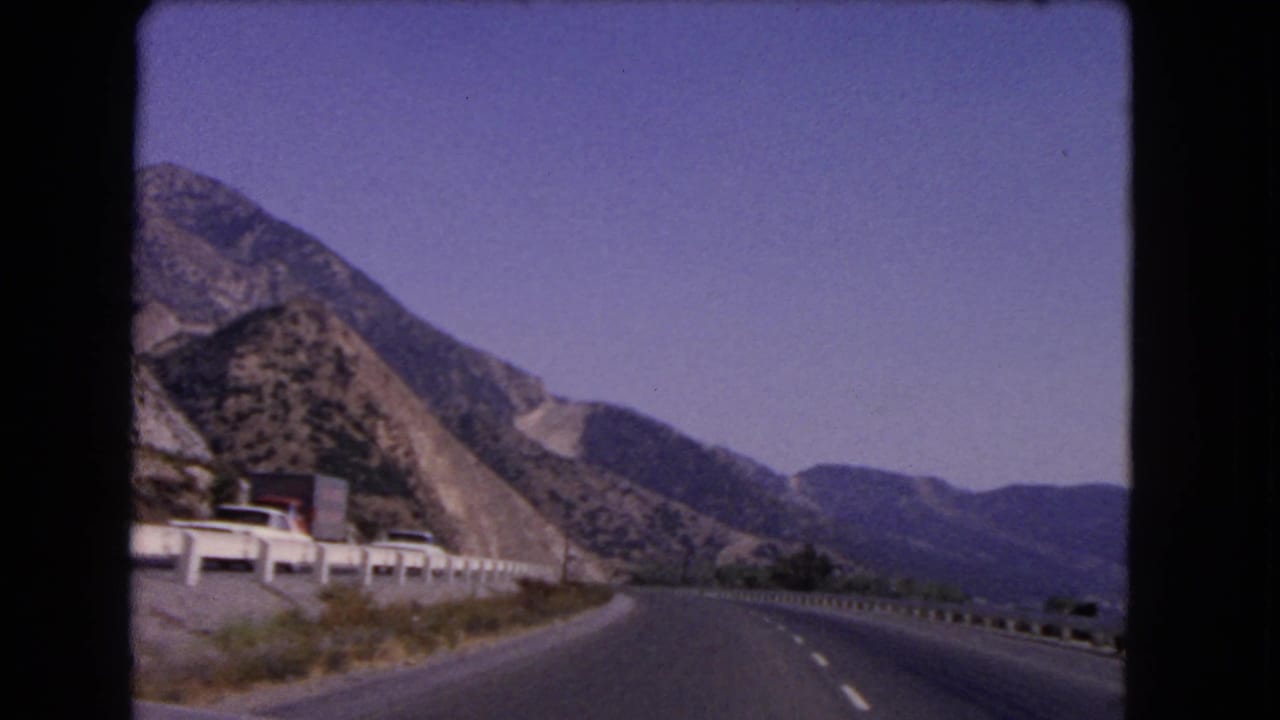 1966 California highway footage shows a bend through rugged mountains with a white guardrail and small distant vehicles.