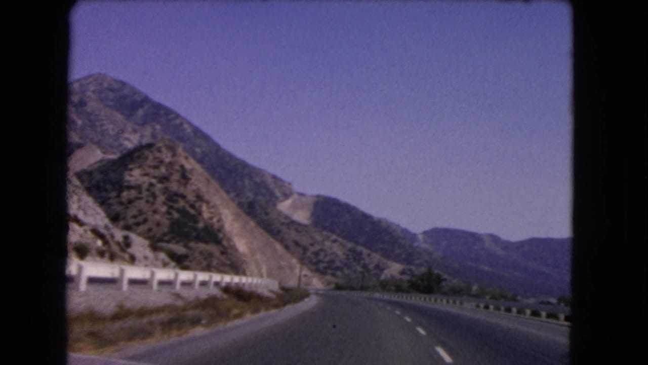 1966 California 8mm frame from a moving vehicle, looking along a mountain road with guardrail, dry slopes, and open sky.