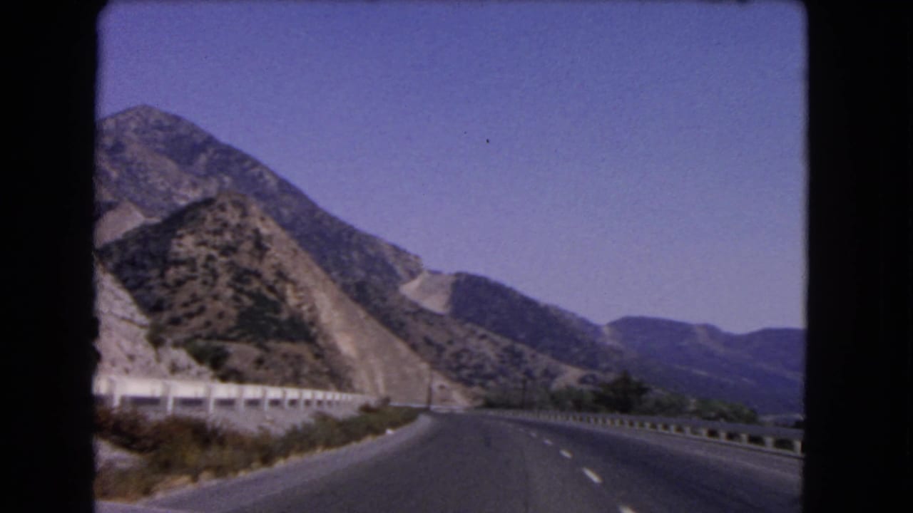 1966 California home-movie frame shows an open mountain highway with guardrails, lane markings, and hazy hills beyond.