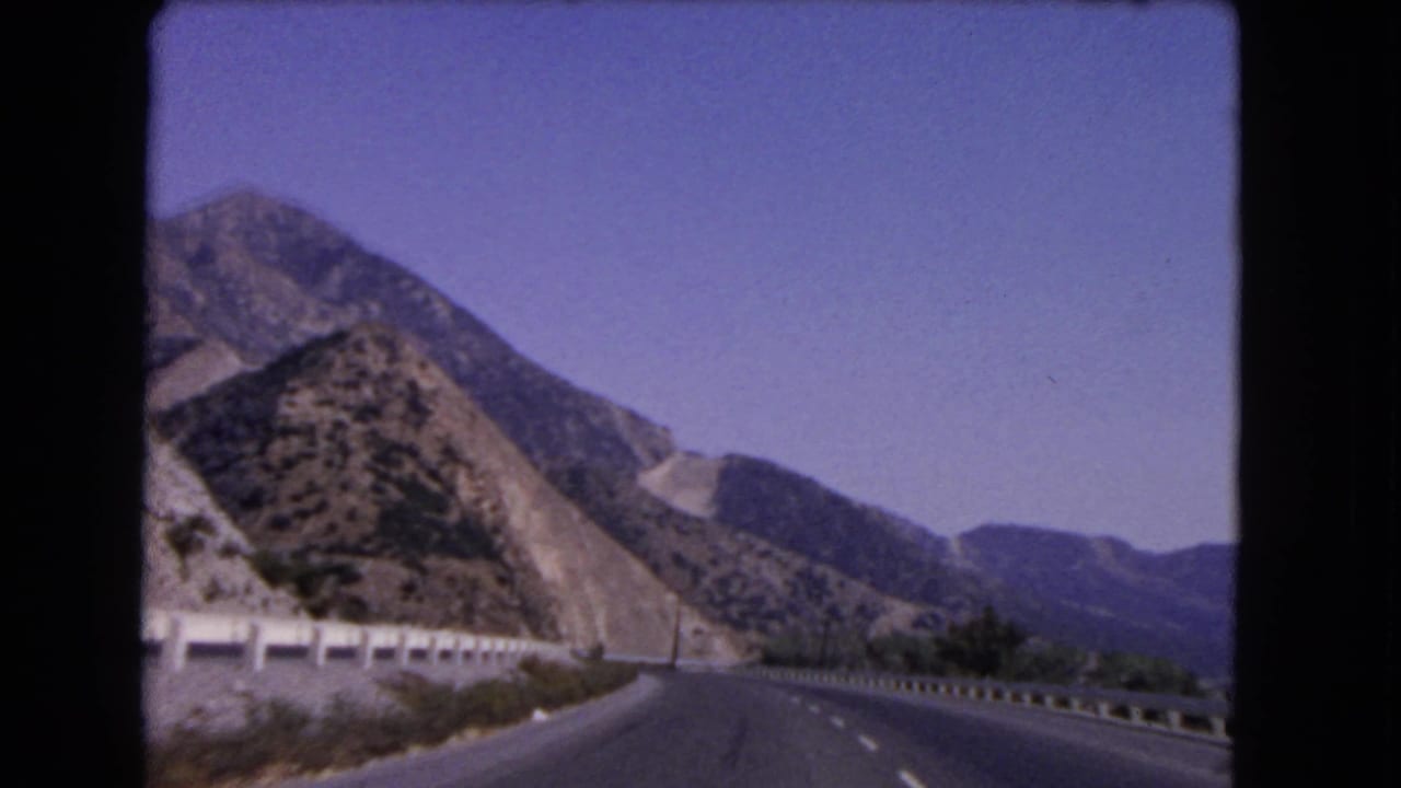 1966 California 8mm frame showing a curving mountain highway, roadside barriers, dry hills, and open blue sky.