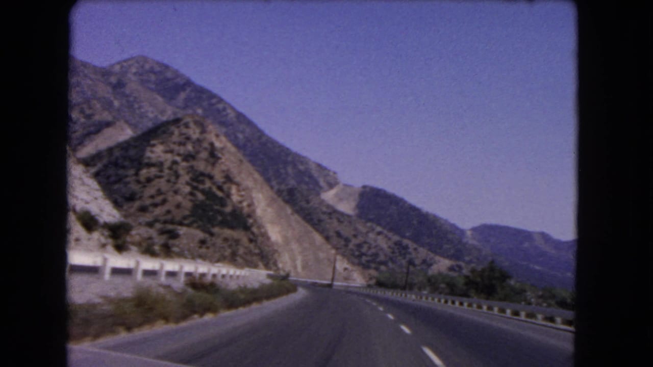 1966 California road-view frame with a divided highway, shoulder barriers, sun-baked hills, and distant mountain ridges.