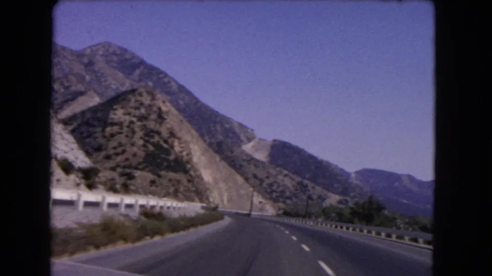 1966 California road-view frame with a divided highway, shoulder barriers, sun-baked hills, and distant mountain ridges.