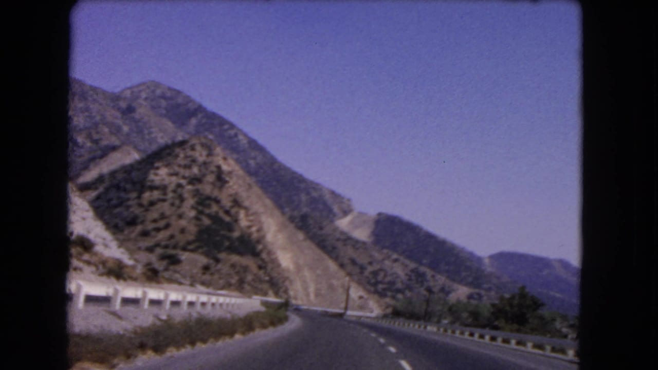 1966 California 8mm view from a moving vehicle on a curving highway bordered by pale guardrails and scrubby hills.