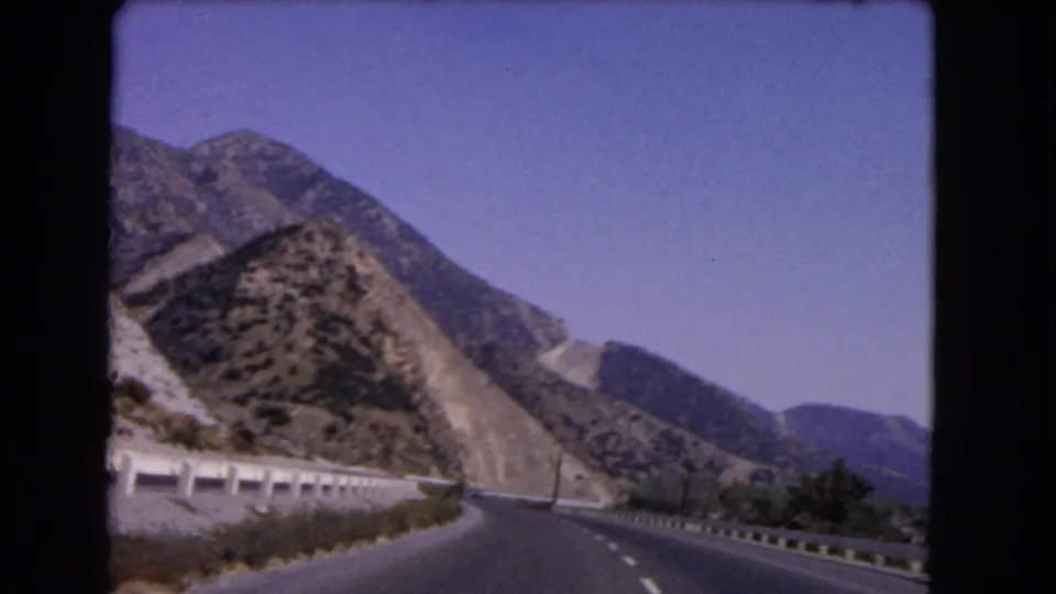 1966 California 8mm view from a moving vehicle on a curving highway bordered by pale guardrails and scrubby hills.