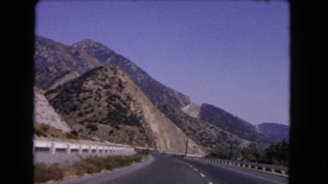 1966 California home-movie frame showing a curving highway below rugged slopes, roadside barriers, and distant hills.