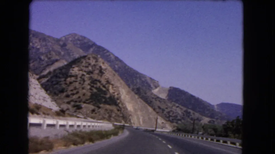 1966 California home-movie frame showing a curving highway below rugged slopes, roadside barriers, and distant hills.