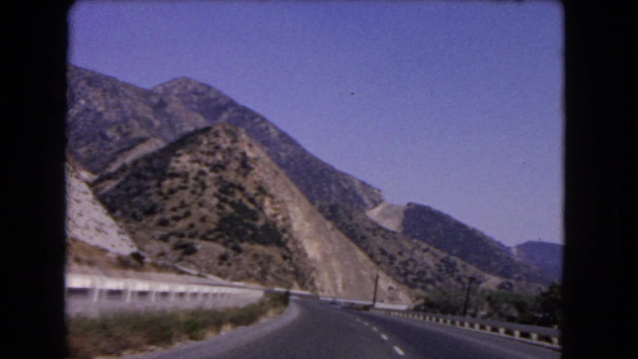 1966 California 8mm frame showing a paved mountain highway curving past dry rocky hills and white guardrails.