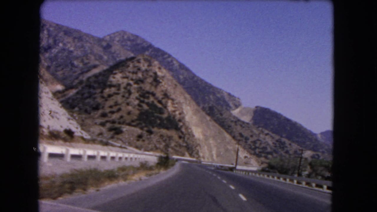 Mountain road curving right with guardrails, utility poles, and rocky slopes beside it.