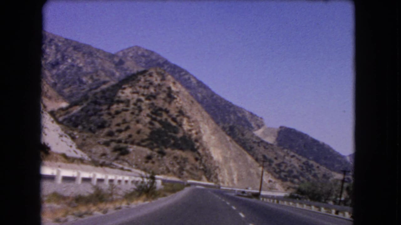 1966 California home-movie frame from a moving car, looking toward a curving highway through rocky hills.