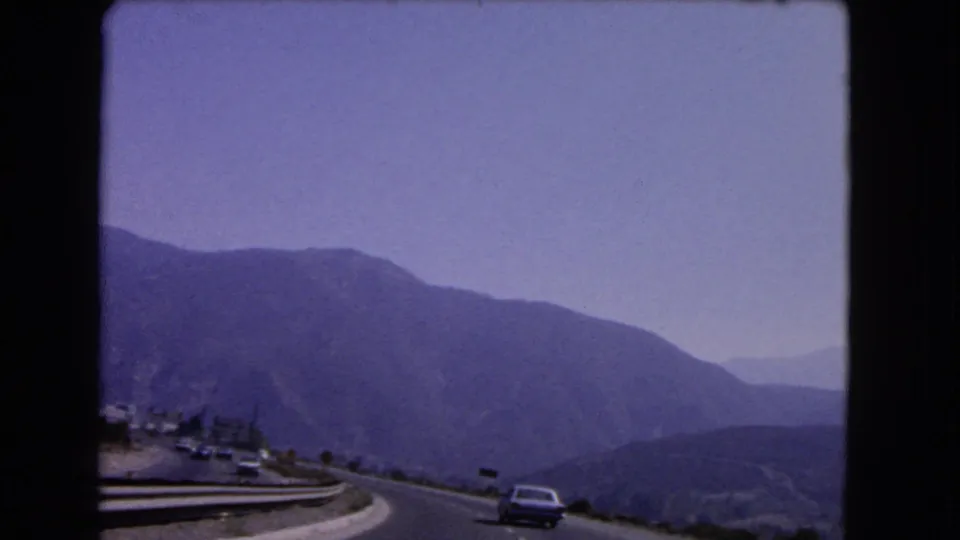 1966 California 8mm frame showing a mountain road with distant cars, guardrails, and hazy layered ridges.