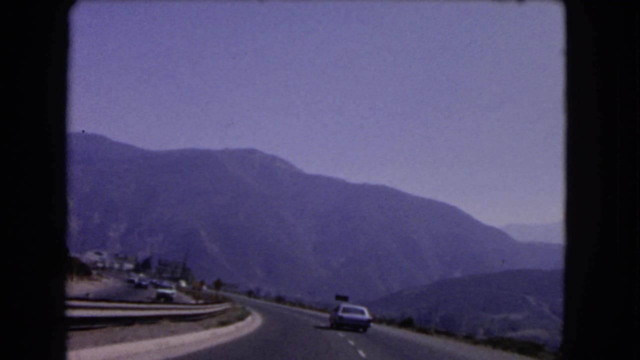1966 California home-movie view from a moving car on a mountain road with a classic car ahead, guardrails, hills, and faded sky.