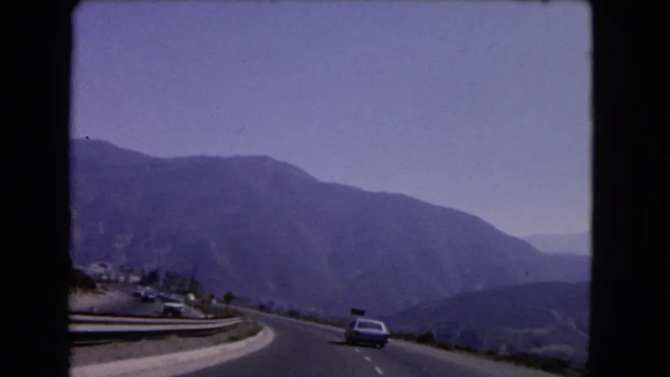 1966 California home-movie view from a moving car on a mountain road with a classic car ahead, guardrails, hills, and faded sky.