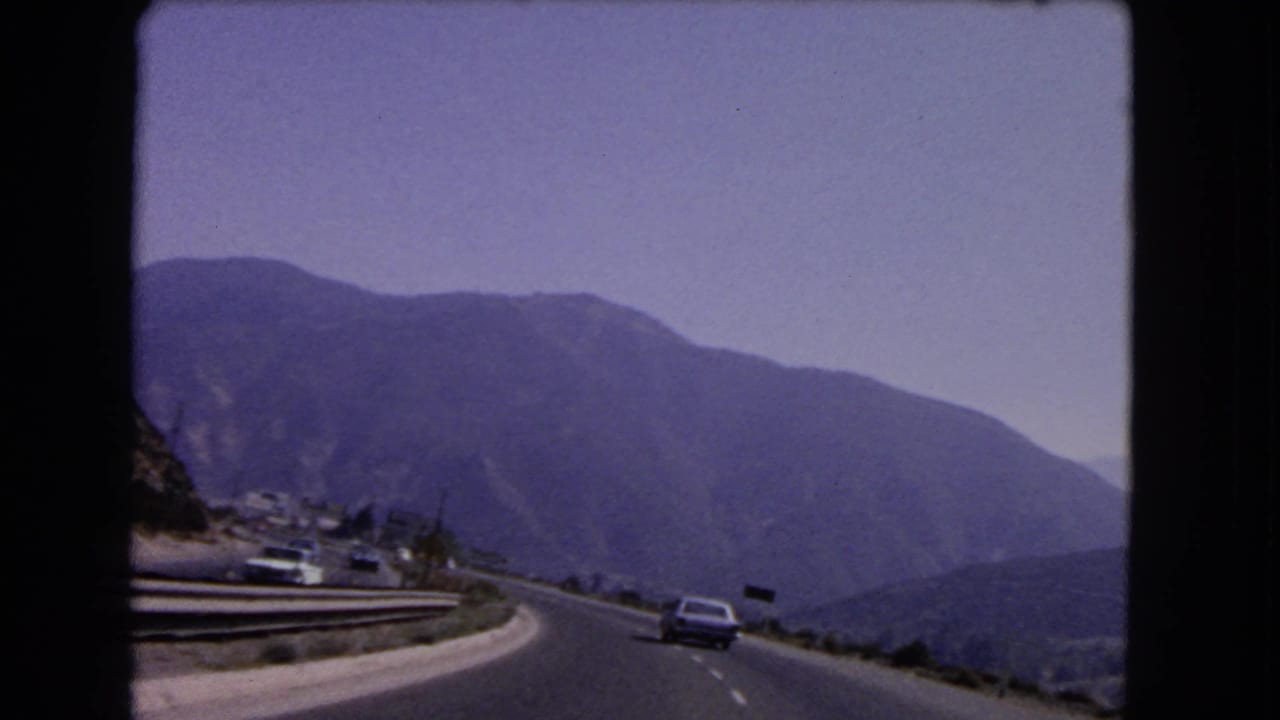 Mountain highway with a car ahead, roadside signs, guardrails, and hazy blue hills.