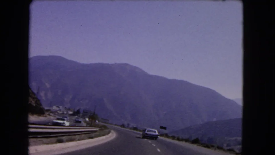 1966 California road scene showing a classic car ahead on a mountain highway with guardrails, small roadside signs, and hazy hills.