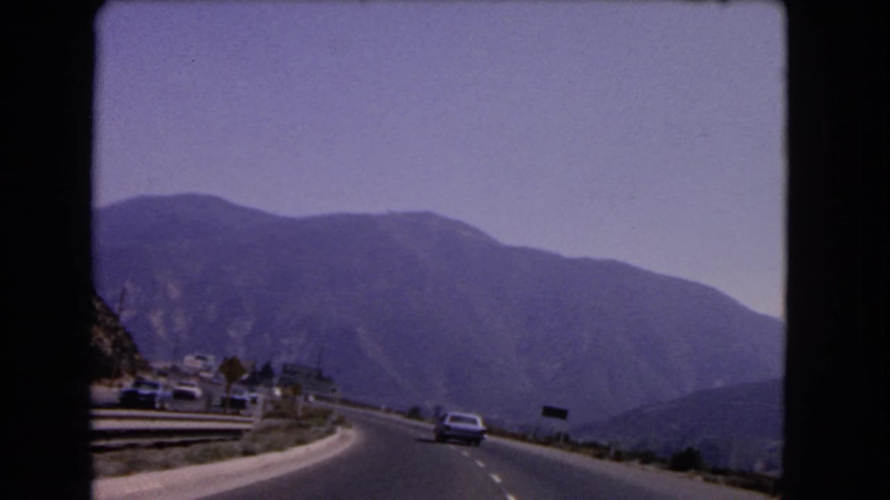 1966 California home-movie frame of a curving mountain road with cars, guardrails, small roadside structures, and broad hills beyond.