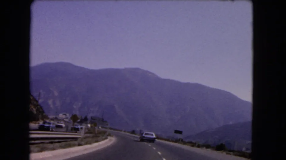 1966 California home-movie frame of a curving mountain road with cars, guardrails, small roadside structures, and broad hills beyond.