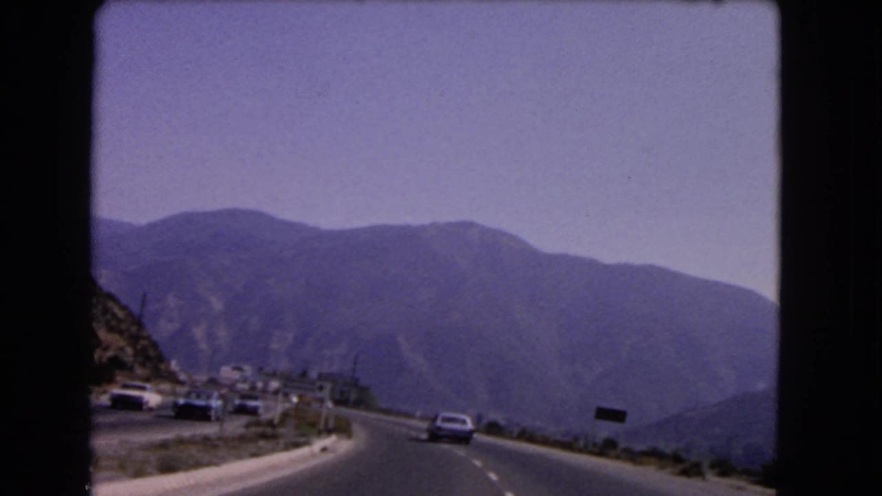 1966 California mountain-road view with a car ahead, a dark roadside sign, guardrails, small structures, and faded blue hills.