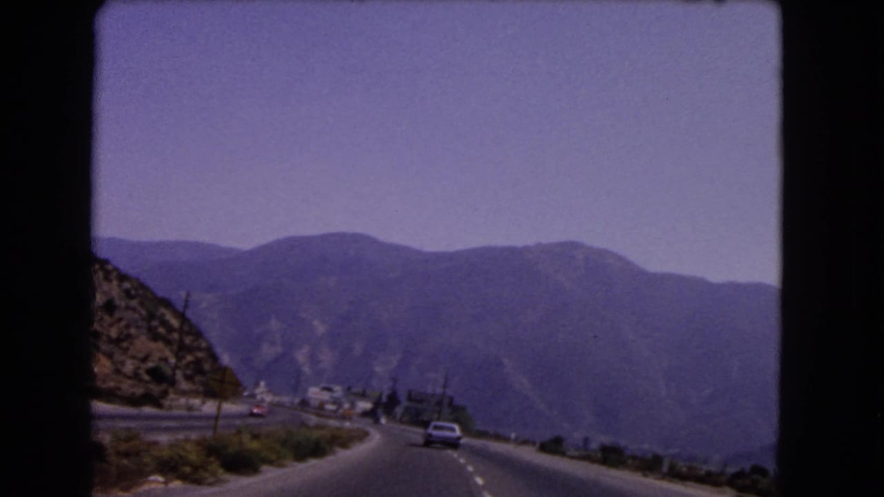 1966 California home-movie frame of a car on a winding mountain highway, filmed from another moving vehicle.