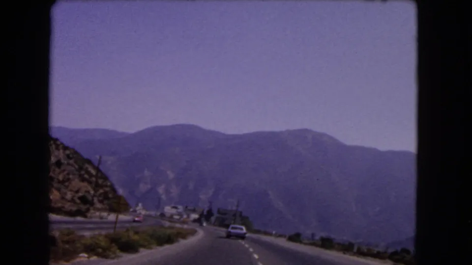 1966 California home-movie frame of a car on a winding mountain highway, filmed from another moving vehicle.